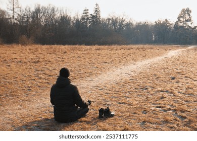 A man sits on dry grass in a field in the cold season and meditates in the lotus position. The man is wearing a black jacket and a black hat, his shoes are nearby - Powered by Shutterstock