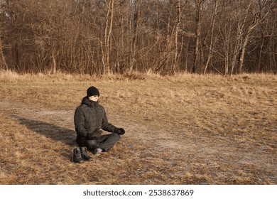 A man sits on dry grass in a field in the cold season and meditates in the lotus position. The man is wearing a black jacket and a black hat, his shoes are nearby - Powered by Shutterstock