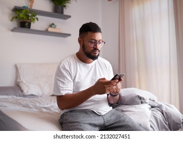 A Man Sits On The End Of His Bed Looking At His Mobile Phone In The Morning. High Quality Photo