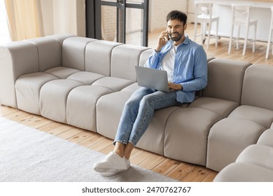 A man sits on a light gray sectional sofa in a modern living room, working on his laptop and talking on his cell phone, working from home - Powered by Shutterstock