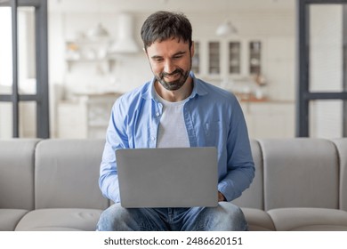 A man sits on a light gray couch in a modern home office, working on his laptop. He is wearing a blue shirt and jeans. He is smiling and looking at the screen. The room is bright and airy. - Powered by Shutterstock