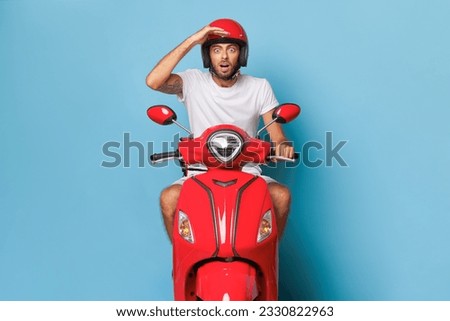 A man sits on red moped in studio, posing isolated on blue background, one hand on his red helmet, another hand on handle bar, fast drive concept, copy space