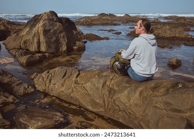 A man sits on a rock by the ocean, wearing a hoodie and jeans, taking a break during a hike. The rocky shoreline and calm waves create a serene and tranquil environment. - Powered by Shutterstock