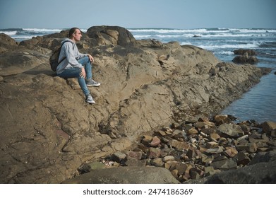 A man sits on a rocky coast, gazing at the ocean waves in the background. He appears peaceful and contemplative, enjoying a moment of tranquility in nature. - Powered by Shutterstock