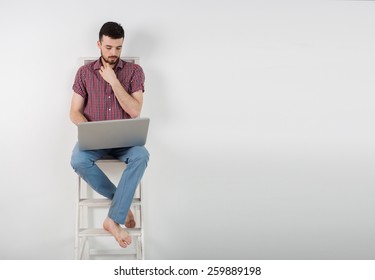 A man sits on a white background ladder on the white wall and working on a laptop. worker wearing a shirt and blue pants - Shutterstock ID 259889198