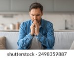 A man sits on a white sofa, his hands clasped together in front of his face as he stares downward, lost in thought. He appears to be pensive and perhaps troubled, as his brow is furrowed.