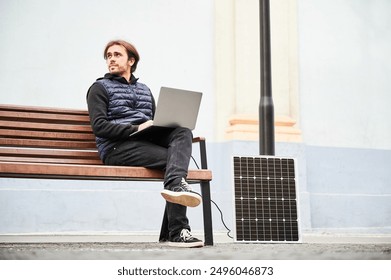 Man sits on wooden bench, working on laptop connected to solar panel. Urban setting and casual attire highlight modern, sustainable, mobile, eco-friendly workspace. - Powered by Shutterstock