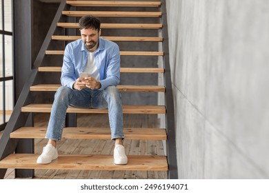 A man sits on a wooden staircase inside a building, smiling and looking at his smartphone. He is wearing blue jeans, a white t-shirt, and a blue button-down shirt. - Powered by Shutterstock