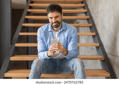 A man sits on wooden stairs inside a home and looks at his smartphone. He is wearing a light blue button-up shirt and denim jeans. The stairs are made of wood and the walls are a light gray color. - Powered by Shutterstock