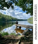 A man sits quietly on a wooden dock, looking out over a calm lake surrounded by trees. The peaceful reflection of the landscape in the water creates a soothing and serene moment