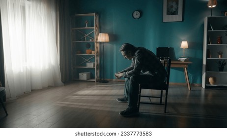 A man sits in the rays of light streaming from a window in a dark room. Side view of a drooping man with his head down, sitting on a chair in the middle of the room. Concept of hope, mental health.
