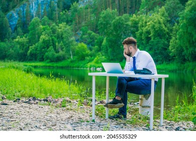 A Man Sits At A Table In Front Of A Laptop And Is Bored In Nature. Office Worker Works At The Computer In The Fresh Air. Businessman At Work.