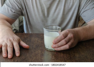 Man Sits At Table With Half-empty Glass Of Kefir. Hand Holds Fermented Milk Drink On Wooden Table. Drink Kefir. Adult Male, Caucasian. Age 44-45. Lifestyle.