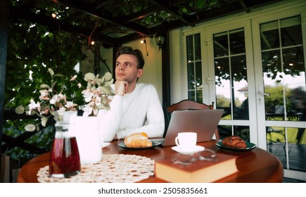 A man sits at a table with a laptop, coffee, and pastries, lost in thought on a beautiful morning. - Powered by Shutterstock