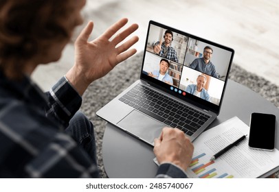 A man sits at a table, participating in a video conference on his laptop. He waves his hand to the camera, engaging with the four other individuals in the video call - Powered by Shutterstock