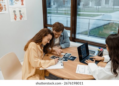 A man sits at a table and talks while pointing at a clipboard on which a smiling pregnant woman is writing, a female doctor is typing on a laptop next to them, in a hospital, during pregnancy