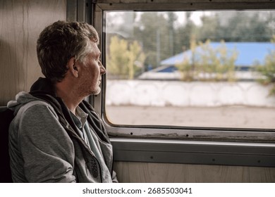 man sits in train, looking thoughtfully out window. he seems deep in thought while traveling through scenic views green fields and distant trees, capturing moment reflection. close up.