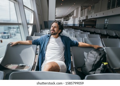 A Man Sits In A Waiting Room At The Gate At The Airport. Young Guy, A Traveler With A Backpack Waiting For The Departure Of The Plane On The Background Of A Large Window And The Runway