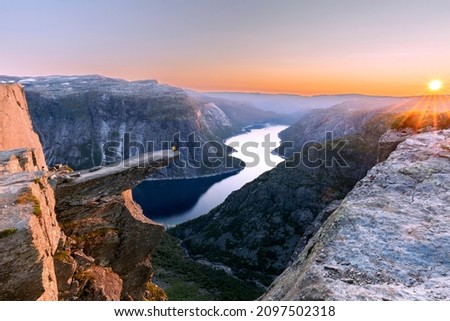 A man sits while throwing his arms in the air on the mountain's cliff edge of Trolltunga throning over Ringedalsvatnet watching the sunset in the snowy Norwegian mountains near Odda, Rogaland, Norway