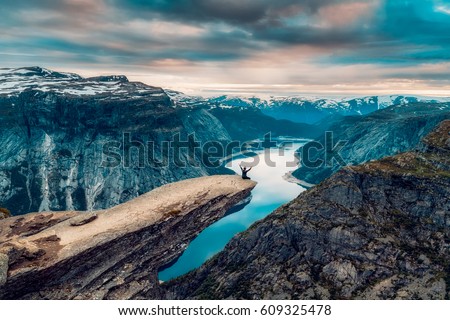A man sits while throwing his arms in the air on the mountain's cliff edge of Trolltunga throning over Ringedalsvatnet watching the sunset in the snowy Norwegian mountains near Odda, Rogaland, Norway.