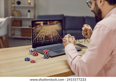 Man sitting back at the desk and playing gambling betting on the internet in online casino at home. Guy with poker chips looking at the screen of his modern laptop computer holding fists for win.