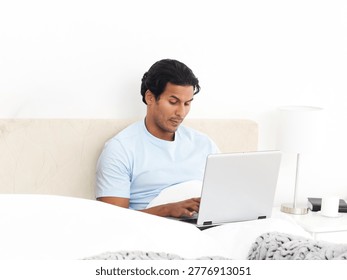 Man sitting in bed using silver laptop, wearing light blue T-shirt, beige headboard behind. Homeoffice, remote, productivity, focus, minimalist, cozy, welllit