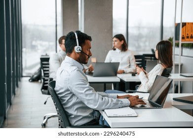 Man is sitting by laptop in headphones. Four people are working in the office together.