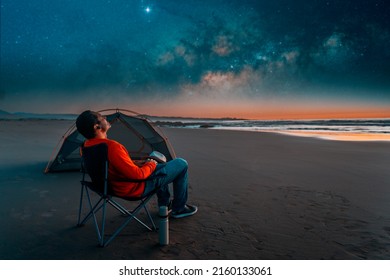 Man Sitting In A Camping Chair Alone On The Beach Looking At The Starry Night Next To A Tent