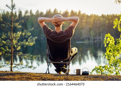 A man is sitting in a camping chair on a summer evening on the background of a forest lake. Object in focus, background blurred.