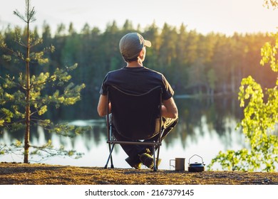 A man is sitting in a camping chair on a summer evening on the background of a forest lake. - Shutterstock ID 2167379145