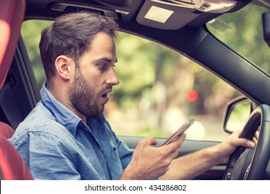 Man Sitting In Car With Mobile Phone In Hand Texting While Driving. Distracted Shocked Guy Checking His Smart Phone Not Paying Attention At Road Annoyed By Bad Text Message Email Outdoors Background