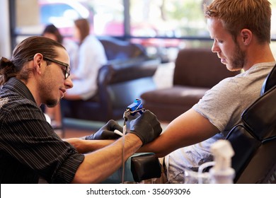 Man Sitting In Chair Having Tattoo On Arm In Parlor