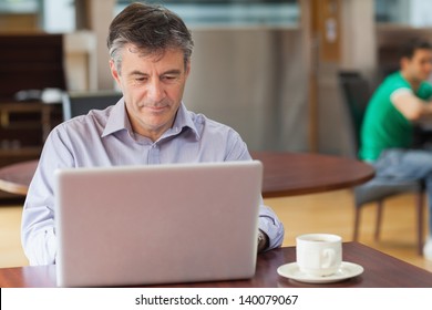 Man Sitting In A Coffee Shop While Drinking A Cup Of Coffee And Using A Laptop