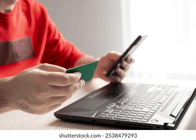 A man is sitting at a desk in a bright room holding a phone and a credit card in front of a laptop, preparing to pay for a purchase. Close-up - Powered by Shutterstock