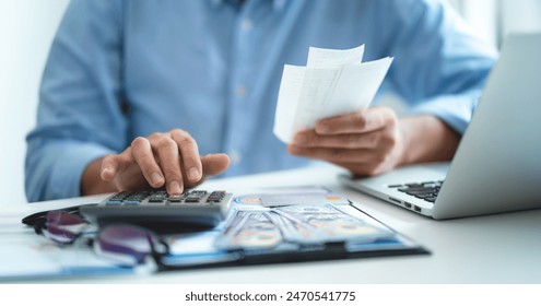 A man is sitting at a desk with a calculator and a stack of bills. He is calculating the total amount of money he has spent