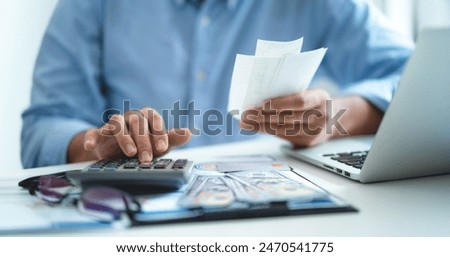 A man is sitting at a desk with a calculator and a stack of bills. He is calculating the total amount of money he has spent