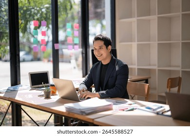 A man is sitting at a desk with a laptop and a stack of papers. He is smiling and he is happy