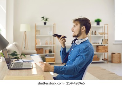 Man Sitting At Desk With Notebook PC In Home Office, Holding Mobile Phone And Talking On Speakerphone. Young Student Or Entrepreneur Recording Voice Message On Messenger Or Using Audio Note Taking App