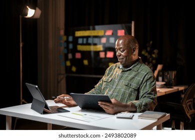 A man is sitting at a desk with two laptops and a tablet. He is looking at the screen and he is focused on his work. The room is dimly lit, which creates a serious and focused atmosphere - Powered by Shutterstock