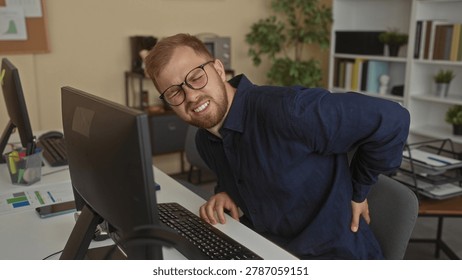 Man sitting at desk using computer with hand on lower back for backache in office building; work stress pain.