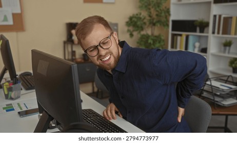 Man sitting at desk using computer with hand on lower back for backache in office building; work stress pain.