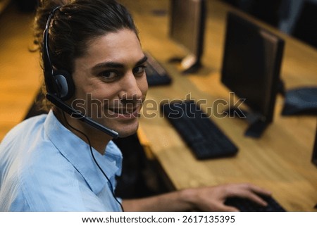 Man sitting at desk using computer and headset, smiling at camera in office setting, copy space. Workstation, call center, helpdesk, customer service, business casual, productivity, approachability