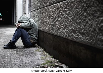 A Man Sitting Down Depressed In A Dark Alleyway At Night With His Head Buried In His Arms