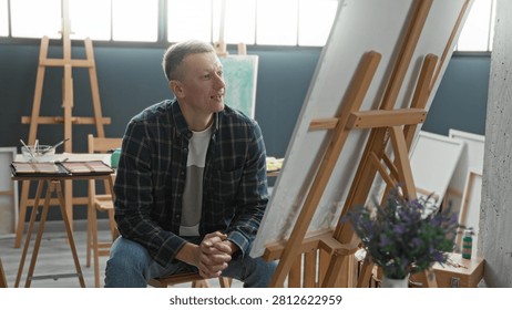 Man sitting at easel, hands clasped on knees, looking intently at canvas amid paint tools, palettes and stacked canvases in studio; creative contemplation.