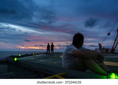 Man Sitting At The Edge Of Jack Up Drilling Rig While Others Walking On Helipad With Helipad's Light On At Sunset Time