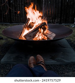 man sitting in front of fire pit (jeans, pants, shoes) looking down at burning campfire (winter outdoor activity)