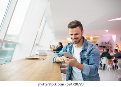 Man is sitting in front of the table near window. He is looking at cellphone and chatting with friends. Smiling young student has coffee break - Powered by Shutterstock