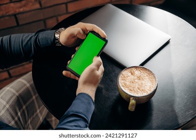 Man sitting and holding smartphone with green screen in cafe with coffee latte and laptop. Freelancer and remote work - Powered by Shutterstock