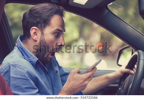 Man\
sitting inside car with mobile phone in hand texting while driving.\
Shocked guy checking his smartphone not paying attention at road\
stunned by bad text message email outdoors\
background