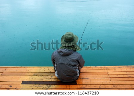 Man sitting at jetty fishing near lake during rainy day